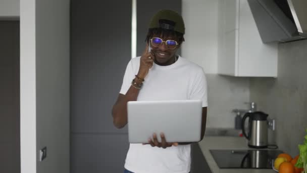アフリカ系アメリカ人の男性が自宅のキッチンでスマートフォンやノートパソコンを使って — ストック動画