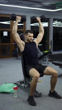 Genç, çekici, kaslı bir sporcu, modern bir spor salonunda dambıllarıyla çalışıyor..