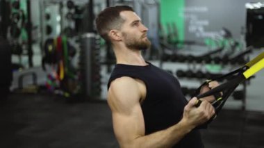Spor salonunda TRX kayışlarıyla egzersiz yapan güçlü sporcunun yan görünümü.