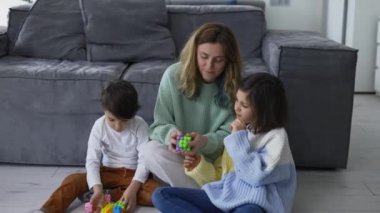 Happy young adult mother playing with two cute small kids, having fun sit on floor.