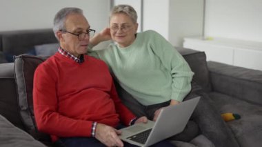 Mature couple looking at laptop, discussing news sitting on sofa.