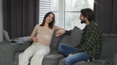 Woman touching pregnant belly, sitting with friend or husband on sofa.