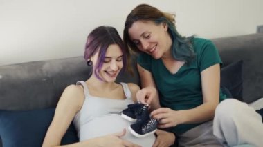 Pregnant lesbian couple sitting on sofa with a pair of baby shoes.