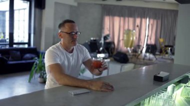 an adult man in glasses sits at the bar counter and takes a sip of a red-colored cocktail.