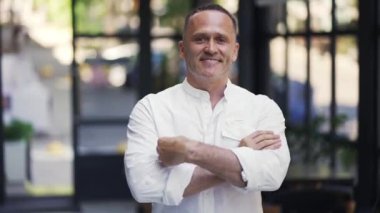 An adult man in a white shirt crosses his arms on his chest against the backdrop of a restaurant.