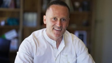 An adult man with stubble in a white shirt sitts in a cafe smiling and posing for the camera.