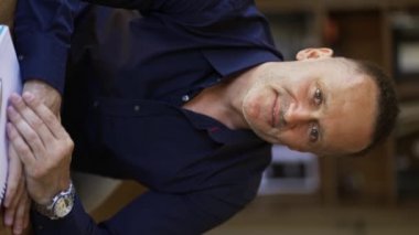 An adult man with stubble in a black shirt sitts in a cafe smiling and posing for the camera.