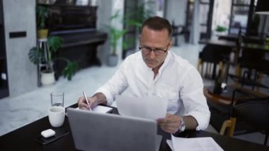 Focused adult businessman working on project, writing paper notes, sitting at caffee desk. Concentrated male professional analyzing market research information.