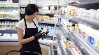 Asian girl worker in an apron with a badge checks the goods on the shelves. Re-inventory of goods, checking the expiration date of goods.