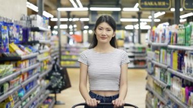 Happy Asian girl walks around the supermarket with a shopping cart and looking at the camera. Shopping trip. daily business.