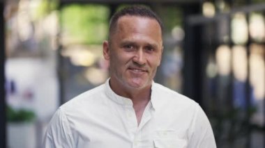 An adult man with stubble in a white shirt stands in a cafe smiling and posing for the camera.