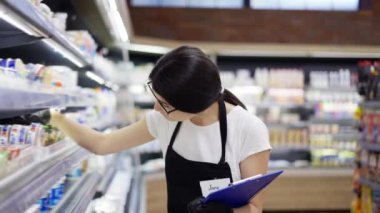 Asian girl worker in an apron with a badge checks the goods on the shelves. Re-inventory of goods, checking the expiration date of goods.