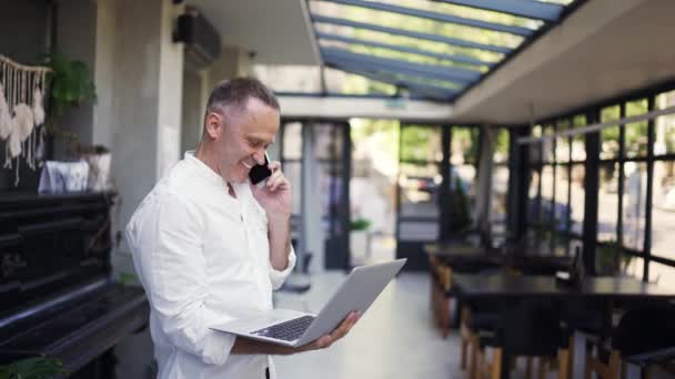 一个穿着衬衫的成年人拿着笔记本电脑走进一家餐馆 在电话里说着话 — 图库视频影像