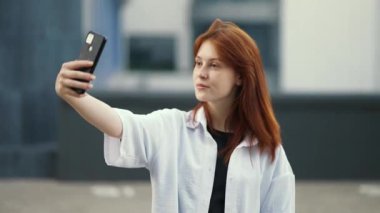 Gömlekli kızıl saçlı kız şehrin arka planında selfie çekiyor..