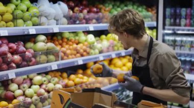 Siyah önlüklü ve eldivenli erkek işçiler süpermarkette meyve stokluyor. İşte genç bir çalışan. Kenar.