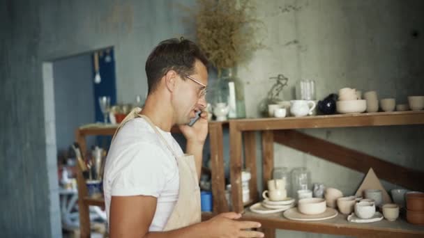 一个戴着眼镜的快乐的年轻陶工站在自己的陶土旁边 一边用智能手机聊天 传统商业概念 — 图库视频影像