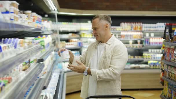 食料品店の中年の訪問者は乳製品を選択します 手にカートを持った男が店内を歩いている 食料品店のショッピングコンセプト — ストック動画