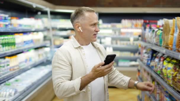 中年の男がビデオリンクで話している食料品店を歩いている 耳の中のヘッドフォン 買い物旅行 現代のコミュニケーション — ストック動画