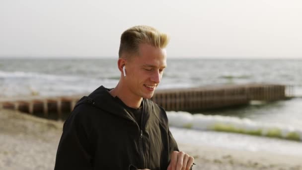 ヘッドフォンで音楽を聴き 午前中にビーチ沿いを歩く若い男の肖像画 — ストック動画