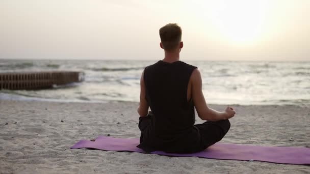 若い男が太陽の夜明けの間に座っている間 ヨガやスポーツマットで瞑想をしています バックビュー 特定の練習をする レクリエーション — ストック動画