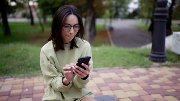 一个身穿绿色毛衣 戴着眼镜的黑发女孩坐在公园的滑板上 在电话里发短信 业余爱好 — 图库视频影像