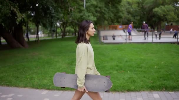 一个女孩手里拿着滑板在公园里散步 那是她每天的嗜好 在公园里散步 带着滑板去滑板公园 — 图库视频影像