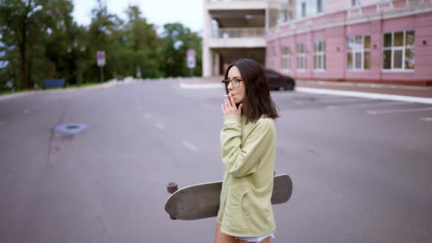 彼女の手にスケートボードと一緒に道路に沿って歩いている緑のセーターや眼鏡でブルネットの肖像画やタバコを吸っています 悪い習慣 — ストック動画