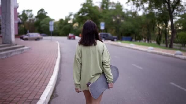 一个穿着绿色毛衣的快乐的黑发姑娘带着滑板在公园里散步 后视镜走在公园里 业余爱好 — 图库视频影像