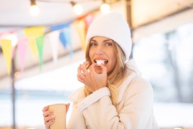 Güzel, mutlu, genç bir kadın lokumla sıcak içecek içiyor. Güzel bir kadın, şehirde kışın elinde kağıt bardak tutuyor.