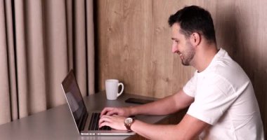 Dizüstü bilgisayarla çalışan genç adamın yan görüntüsü, zamanı kontrol etmek ve gece evde kalan işleri bitirmek.