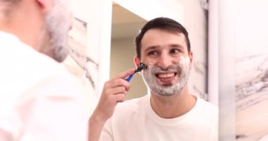 Yakışıklı, gülümseyen genç bir adam tıraş olurken ve banyoda akıllı telefonla konuşurken.