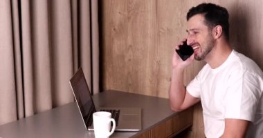 Genç adam dizüstü bilgisayar kullanıyor, akıllı telefonla konuşuyor ve evdeki beyaz bardaktan içiyor.