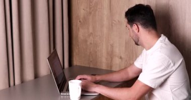 Bilgisayarı kullanan, akıllı telefonla konuşan ve evdeki beyaz bardaktan içen gülümseyen genç adamın yan görüntüsü.