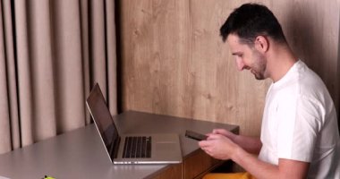 Sırıtkan genç adam dizüstü bilgisayarla masada otururken akıllı telefon kullanıyor.