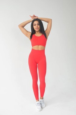 Kırmızı spor giyimli Afro-Amerikan kadın ellerini başının üstüne koyup gri arka planda kameraya gülümsüyor.