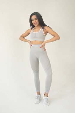 Gri spor kıyafetleri içinde, kameraya gülümseyen ve stüdyoda poz veren güzel bir genç kadın. 