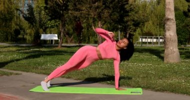 Genç, çok ırklı, pembe spor kıyafetleri, pantolon ve bluz giyen, parkta spor paspası, sağlık sigortası ve zihinsel terapi üzerine yoga egzersizleri yapan çekici bir kadın..