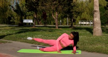 Genç, çok ırklı, pembe spor kıyafetleri, pantolon ve bluz giyen, parkta spor paspası, sağlık sigortası ve zihinsel terapi üzerine yoga egzersizleri yapan çekici bir kadın..