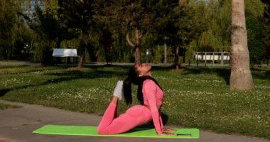 Genç, çok ırklı, pembe spor kıyafetleri, pantolon ve bluz giyen, parkta spor paspası, sağlık sigortası ve zihinsel terapi üzerine yoga egzersizleri yapan çekici bir kadın..