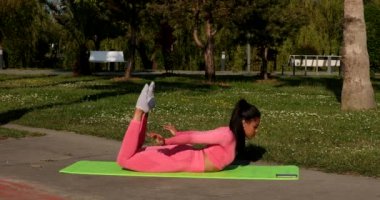 Genç, çok ırklı, pembe spor kıyafetleri, pantolon ve bluz giyen, parkta spor paspası, sağlık sigortası ve zihinsel terapi üzerine yoga egzersizleri yapan çekici bir kadın..
