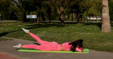 Genç, çok ırklı, pembe spor kıyafetleri, pantolon ve bluz giyen, parkta spor paspası, sağlık sigortası ve zihinsel terapi üzerine yoga egzersizleri yapan çekici bir kadın..