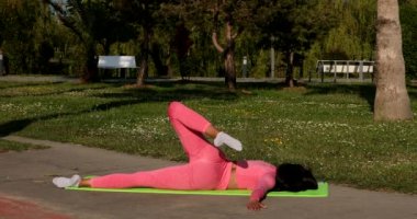 Genç, çok ırklı, pembe spor kıyafetleri, pantolon ve bluz giyen, parkta spor paspası, sağlık sigortası ve zihinsel terapi üzerine yoga egzersizleri yapan çekici bir kadın..