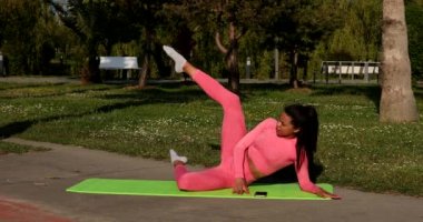 Genç, çok ırklı, pembe spor kıyafetleri, pantolon ve bluz giyen, parkta spor paspası, sağlık sigortası ve zihinsel terapi üzerine yoga egzersizleri yapan çekici bir kadın..