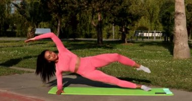 Genç, çok ırklı, pembe spor kıyafetleri, pantolon ve bluz giyen, parkta spor paspası, sağlık sigortası ve zihinsel terapi üzerine yoga egzersizleri yapan çekici bir kadın..