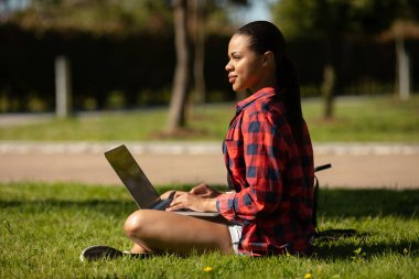 Dışarıda dizüstü bilgisayarı olan neşeli Afro-Amerikan öğrenci. Bilgisayarla çimenlerde oturan, internette sörf yapan ya da sınavlara hazırlanan gülümseyen bir kadın.. 