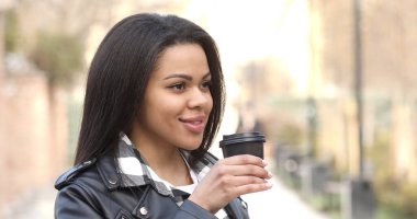 Young black woman outdoors drinking coffee from reusable cup and walking in city.