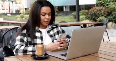 Smiling girl sitting in a street cafe and working at the computer. Young woman checking the data on the laptop and using the phone.