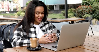 Smiling girl sitting in a street cafe and working at the computer. Young woman checking the data on the laptop and using the phone.