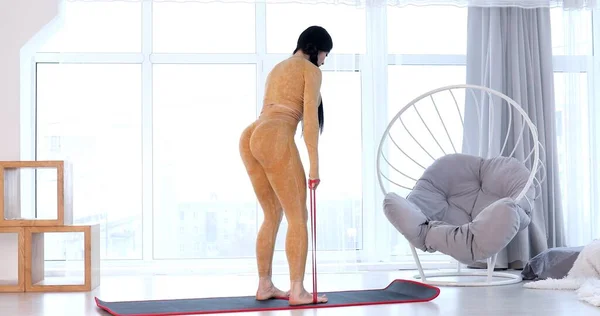 Young fit woman in sportswear doing fitness aerobic exercises with elastic band in living room. Home fitness and wellness concept.