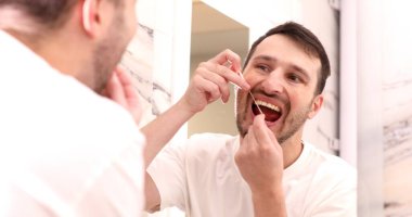 Gülümseyen genç adam aynaya bakıyor ve diş ipliği kullanıyor.